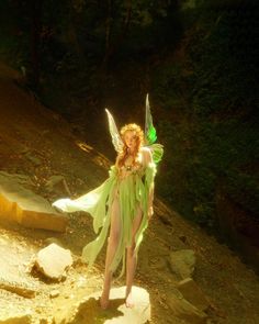 a woman dressed as a fairy walking down a dirt path in the woods with green wings