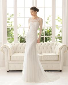 a woman in a white wedding dress standing next to a couch with a window behind her