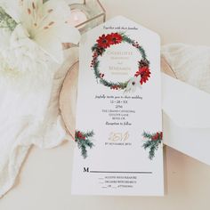 a white wedding card with red flowers and greenery on it next to a candle