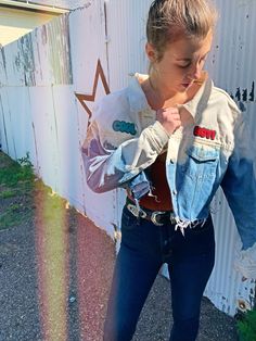 a woman standing in front of a white wall wearing jeans and a jean jacket with patches on it