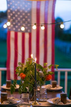 an american flag is in the background behind a table set with plates and cups, candles, and flowers