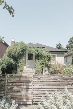 a wooden fence in front of a house with white flowers on the ground and bushes around it