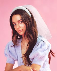 a young woman in a blue sweater and white veil sitting on a pink background with her hands together