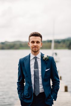 a man in a suit and tie standing on a dock next to the water with his hands in his pockets