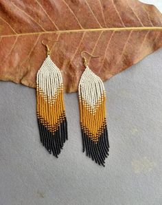 two tone beaded fringe earrings with black and yellow accents on a gray surface next to a leaf