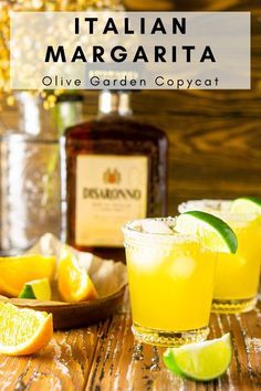 two glasses filled with lemonade and limes on top of a wooden table next to bottles