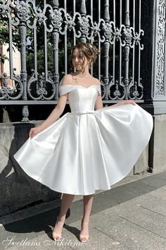 a woman wearing a white dress standing in front of a wrought iron fence with her hands on her hips