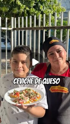 two people standing next to each other holding plates with food on them in front of a fence