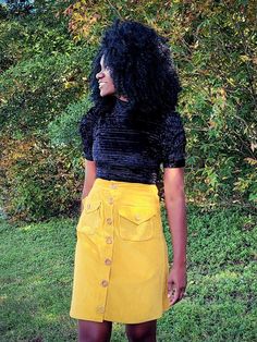 a woman standing in front of some bushes wearing a yellow skirt and black shirt with buttons