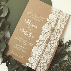 a brown and white wedding card with lace on the front is laying next to greenery