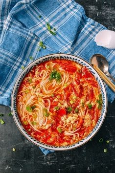 a bowl of soup with noodles, meat and vegetables on a blue towel next to two spoons