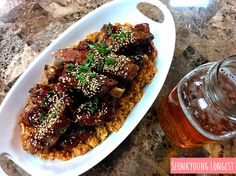 a white plate topped with meat and rice next to a glass of beer on top of a table