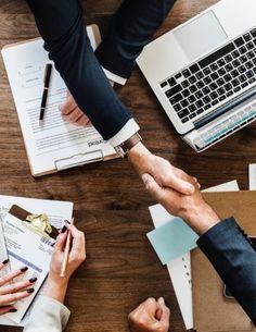 two people shaking hands while sitting at a table in front of laptops and papers