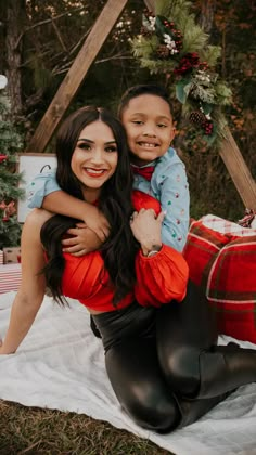 a woman holding a small child in her arms while sitting on a blanket near a christmas tree