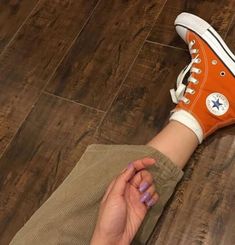 a woman's hand holding onto an orange converse shoe on top of a wooden floor