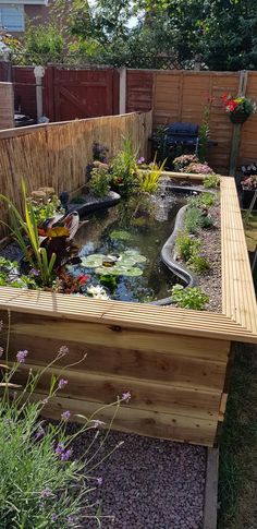 an outdoor pond in the middle of a garden with plants and flowers around it, next to a wooden fence