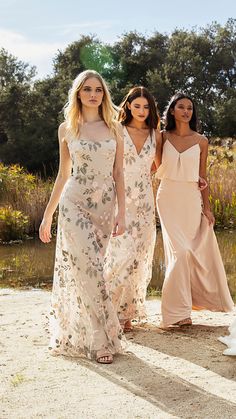 three women in dresses walking on the sand near water with trees and bushes behind them