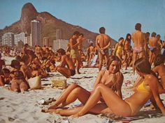 a group of people sitting on top of a sandy beach next to the ocean with mountains in the background