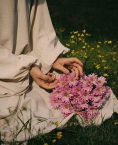 a person sitting in the grass holding a bunch of pink and yellow flowers with their hands