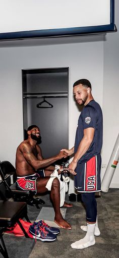 two men standing in a locker room one is tying the other's shoes