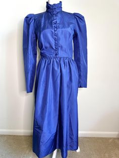 This is a beautiful, special occasion dress from the 1980s by Liz Claiborne. Depending on the light, it can look royal blue or periwinkle.  Ruched high neck, gathered puff sleeves, covered buttons, princess seams and a gathered waist are some of the details that make this dress so pretty. Fitted bodice. The bottom button on the bodice is a replacement and the belt is missing. Three covered buttons on the sleeves. A few discolorations along the hem that really do not show up unless you are looking closely for them because the fabric is sort of iridescent (see last photo). This hasn't been dry cleaned, so they may come out. No odors.  Made in Hong Kong.  FLAT MEASUREMENTS Shoulder to shoulder 13" Shoulder to sleeve tip 24-1/4" Armpit to armpit 17" Shoulder to waist 16.5" Waist to hem 28.5" W Blue Floor-length Taffeta Dresses, Blue Cotton Puff Sleeve Dress With Gathered Sleeves, Blue Taffeta Floor-length Dresses, Blue 1940s Dress, 1970s Blue Long Sleeve Dress, Puffy Sleeves, Princess Seam, Liz Claiborne, Tea Dress