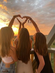 three girls standing on a balcony with their hands in the shape of a heart at sunset