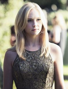 a woman with blonde hair wearing a black dress and looking off into the distance while standing in a park