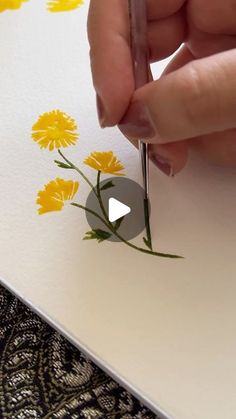 someone is painting flowers on paper with yellow and white colors, while holding a pen in their left hand