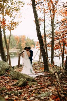 a bride and groom are standing in the woods by the water with their arms around each other