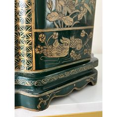 an ornate green and gold painted cabinet with flowers on the front, sitting on a white surface