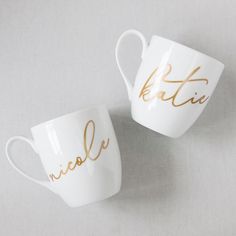 two white cups with gold lettering on them sitting side by side against a gray background