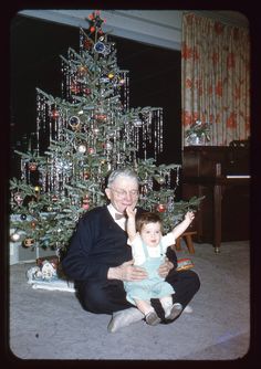 an old man holding a small child in front of a christmas tree