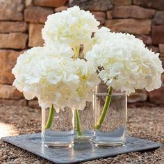 three clear vases with white flowers in them sitting on a stone slab next to a brick wall