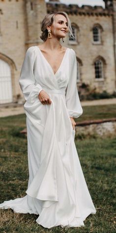 a woman standing in front of a castle wearing a white dress with long sleeves and an asymmetrical neckline