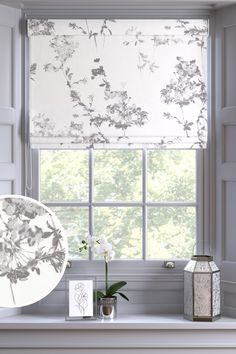 a window with a white flowered roman blind in front of it and an empty vase on the windowsill