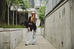 a woman in cowboy hat standing on sidewalk next to wall and building with trees behind her