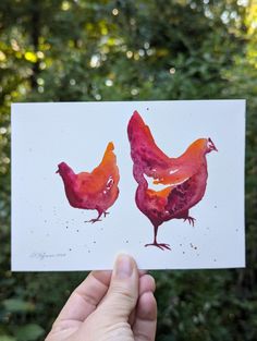 a hand holding up a piece of paper with two chickens painted on it's sides