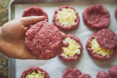 a hand holding a hamburger in front of other burgers on a baking sheet with cheese