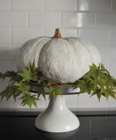 two white pumpkins sitting on top of a cake stand with green leaves around them