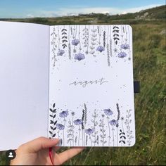 a hand holding an open notebook in front of a field with wildflowers and grass