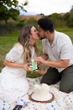 a man and woman kissing while sitting on a blanket with a cake in front of them