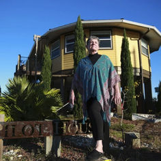 an older woman standing in front of a house with trees and bushes around her feet