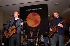 three men are playing guitars in front of a sign that says coldplay parachutes