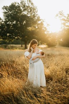 a woman in a white dress holding a baby while standing in tall grass with the sun behind her