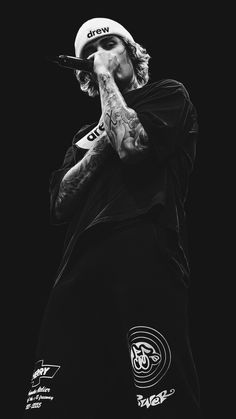 a man with tattoos and a hat on holding his hand up to his mouth while standing in front of a black background