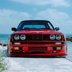 the front end of a red car parked on top of a sandy beach next to the ocean