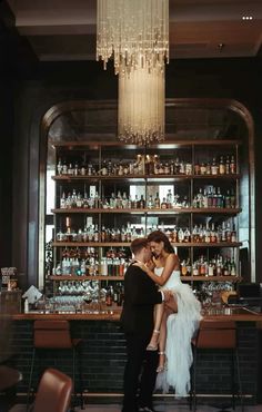 a bride and groom kissing in front of a bar with liquor bottles on the shelves