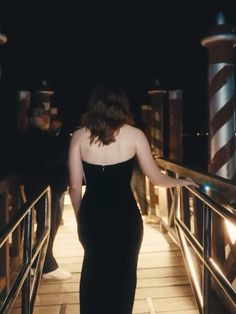 a woman in a black dress is walking down a bridge with her hand on the railing