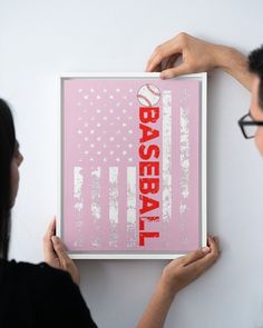 a woman holding up a framed baseball poster with an american flag on the wall behind her