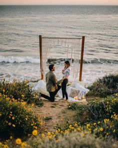 two people sitting on the ground next to an open air hammock by the ocean